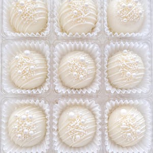Op de afbeelding: Negen met witte chocolade bedekte lekkernijen in een doorzichtige plastic container. Elke ronde lekkernij is versierd met witte drizzle, parelkleurige sprinkles en witte suikerkristallen. De lekkernijen zitten in witte papieren cupcakevormpjes.