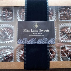 Pu&ograve; includere: Una scatola di nove tartufi al cioccolato con vortici bianchi e marroni. La scatola &egrave; etichettata "Bliss Lane Sweets" con un numero di telefono e un indirizzo email.