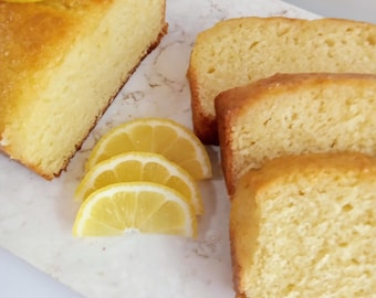May include: A golden-brown lemon loaf cake with a slice of lemon on top and three slices of cake. The cake is on a white marble serving board with several lemon slices. The cake is a dessert.