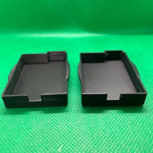 Könnte beinhalten: Zwei schwarze rechteckige Plastikboxen mit einer strukturierten Oberfläche. Die Boxen sind offen und leer.