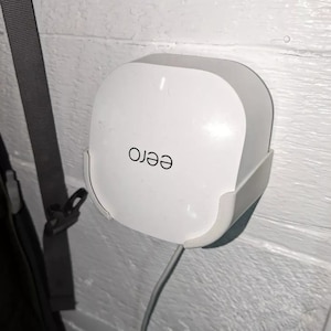 Op de afbeelding: Een witte eero Wi-Fi-router gemonteerd op een witte muur met een wit snoer erin gestoken.