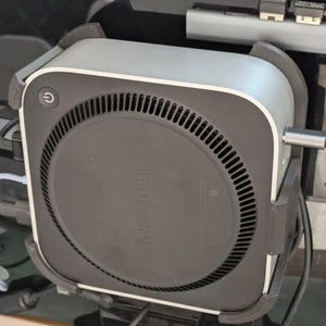 以下が含まれることがあります： 黒い円形の通気孔と「Mac mini」の文字が底に印刷された、シルバーとグレーのMac Miniコンピュータ。 コンピュータは黒いブラケットに取り付けられています。
