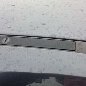 May include: A close-up of a dark gray rectangular object with a textured surface and a circular detail. The object is set against a light gray background with water droplets.