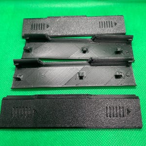 Puede incluir: Cuatro piezas de plástico negro con una superficie texturizada y pequeños agujeros. Las piezas están apiladas una encima de la otra.
