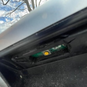 May include: Close-up of a car's rear bumper with a small, yellow button visible inside a black compartment.