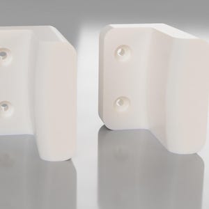 Könnte beinhalten: Zwei weiße Wandhalterungen aus Kunststoff mit Schraubenlöchern. Die Halterungen sind L-förmig und haben eine abgerundete Kante.