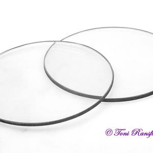 Könnte beinhalten: Zwei klare, kreisförmige Glaslinsen mit dunkelgrauen Rändern überlappen sich auf weißem Hintergrund. Die Linsen scheinen für Brillen oder ein ähnliches optisches Gerät bestimmt zu sein. Das Bild ist gut beleuchtet und die Linsen sind scharf.