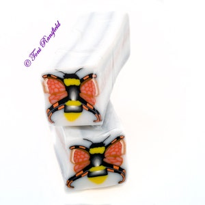 Puede incluir: Dos canes de arcilla polimérica blanca con un diseño de abeja negra y amarilla con alas rojas. La abeja está centrada en cada cane.