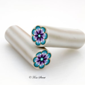 Puede incluir: Dos bastones de arcilla polimérica con diseños florales. Los extremos de los bastones muestran una flor con detalles turquesa, morado y dorado. Los bastones son de color gris claro y cilíndricos, sobre una superficie blanca. La imagen está bien iluminada.