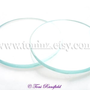 Peut inclure: Deux lentilles en verre transparentes et circulaires sont présentées sur un fond blanc. Les lentilles ont une légère teinte bleu-vert sur les bords. Le texte "www.tonimz.etsy.com" est visible en arrière-plan. L'image est signée "© Toni Ransfield".