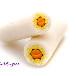 May include: Two white polymer clay canes with yellow and orange flower designs. The canes are rolled and ready to be used for crafting.