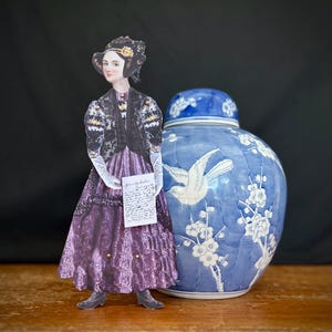 May include: A paper doll of a woman in a purple dress and black jacket holding a letter stands next to a blue and white porcelain ginger jar with a bird and floral design.