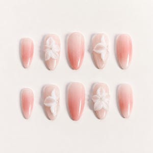 May include: A set of ten artificial nails in a soft pink gradient. Some nails feature a 3D white flower design. The nails are oval-shaped and arranged on a white surface, offering a delicate and feminine aesthetic.