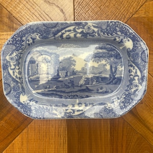 May include: A blue and white ceramic serving platter with a rectangular shape. The centre features a landscape scene, surrounded by a floral border. The platter's dimensions are approximately 30 cm in length and 20 cm in width.