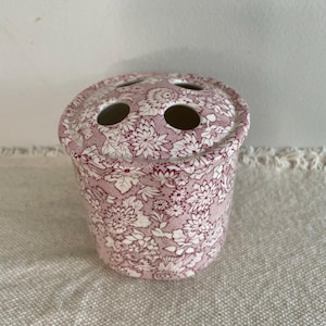 May include: A pink and white floral toothbrush holder. The ceramic holder has three holes on the top and a cylindrical shape. The design features a repeating pattern of flowers, and is a classic, vintage style.