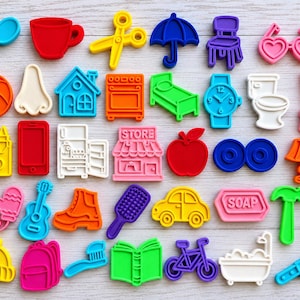 May include: A collection of colorful plastic cookie cutters in various shapes including a spoon, cup, scissors, umbrella, chair, heart glasses, ball, nose, house, oven, bed, watch, toilet, broom, crayons, phone, fridge, store, apple, lamp, mittens, guitar, boot, brush, car, soap, hammer, key, hat, backpack, toothbrush, book, bicycle, bathtub, and knife.
