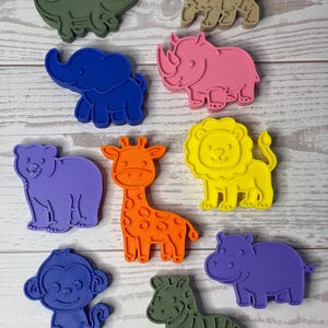 May include: A collection of ten plastic animal-shaped cookie cutters in various colors, including a green crocodile, tan tiger, blue elephant, pink rhinoceros, purple bear, orange giraffe, yellow lion, blue monkey, green zebra, and purple hippo.