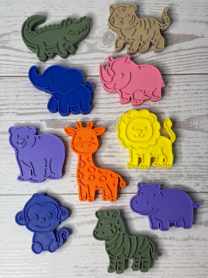 May include: A collection of ten plastic animal-shaped cookie cutters in various colors, including a green crocodile, tan tiger, blue elephant, pink rhinoceros, purple bear, orange giraffe, yellow lion, blue monkey, green zebra, and purple hippo.