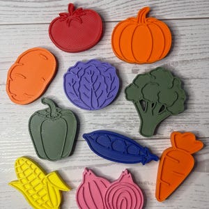 May include: A collection of ten plastic vegetable and fruit shaped cookie cutters in various colors including red, orange, purple, green, blue, yellow, and pink. The set features shapes of a tomato, pumpkin, potato, cabbage, broccoli, pepper, pea pod, corn, onion, and carrot.