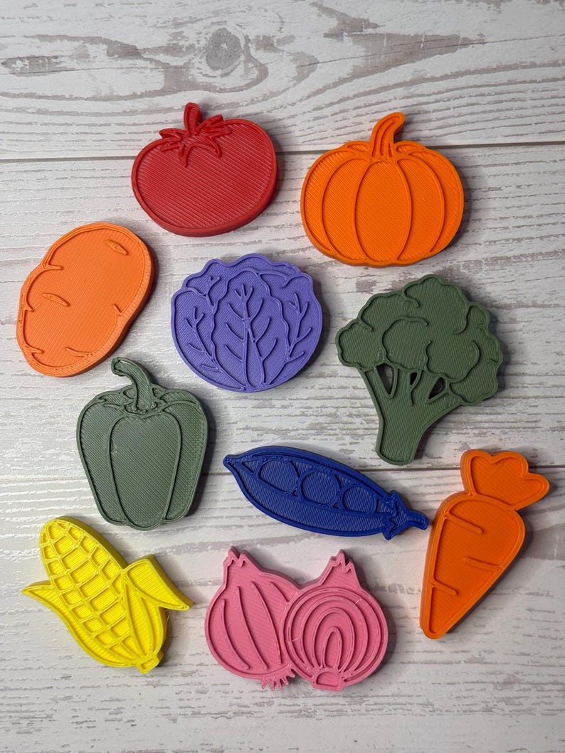 May include: A collection of ten plastic vegetable and fruit shaped cookie cutters in various colors including red, orange, purple, green, blue, yellow, and pink. The set features shapes of a tomato, pumpkin, potato, cabbage, broccoli, pepper, pea pod, corn, onion, and carrot.