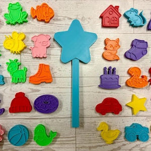 May include: A collection of colorful, plastic cookie cutters in various shapes, including animals, objects, and a star-shaped wand. The cutters are in shades of pink, green, orange, yellow, blue, and purple. The shapes include a dog, frog, fish, house, dress, and more.