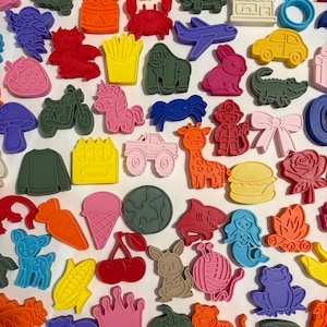 May include: A collection of colorful, rubbery charms in various shapes, including animals, food items, and vehicles. The charms are in shades of pink, green, yellow, and blue, with a few in orange and red. The assortment includes a unicorn, a car, and a hamburger.