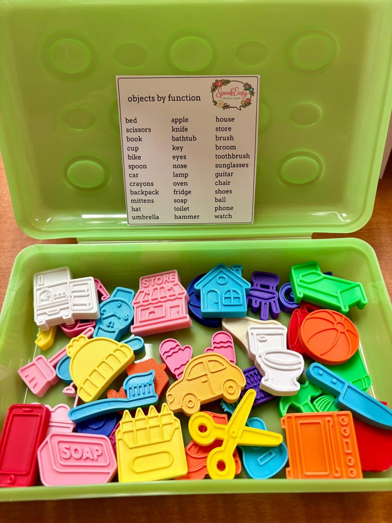 May include: A lime green plastic storage container filled with various colorful, small, flat plastic shapes representing objects like a car, house, scissors, and soap. A white label inside the lid lists the objects by function in black text.