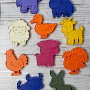May include: A collection of ten plastic animal-shaped cookie cutters in various colors including purple, blue, white, orange, yellow, pink, and green. The set features a horse, cow, sheep, duck, goat, chicken, pig, turkey, bull, and donkey.