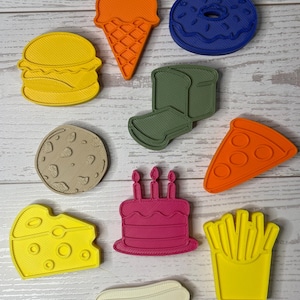 May include: A collection of ten plastic food-themed cookie cutters in various colors, including a yellow burger, orange ice cream cone, blue donut, green sandwich, tan cookie, orange pizza slice, yellow cheese wedge, pink birthday cake, and white hot dog.