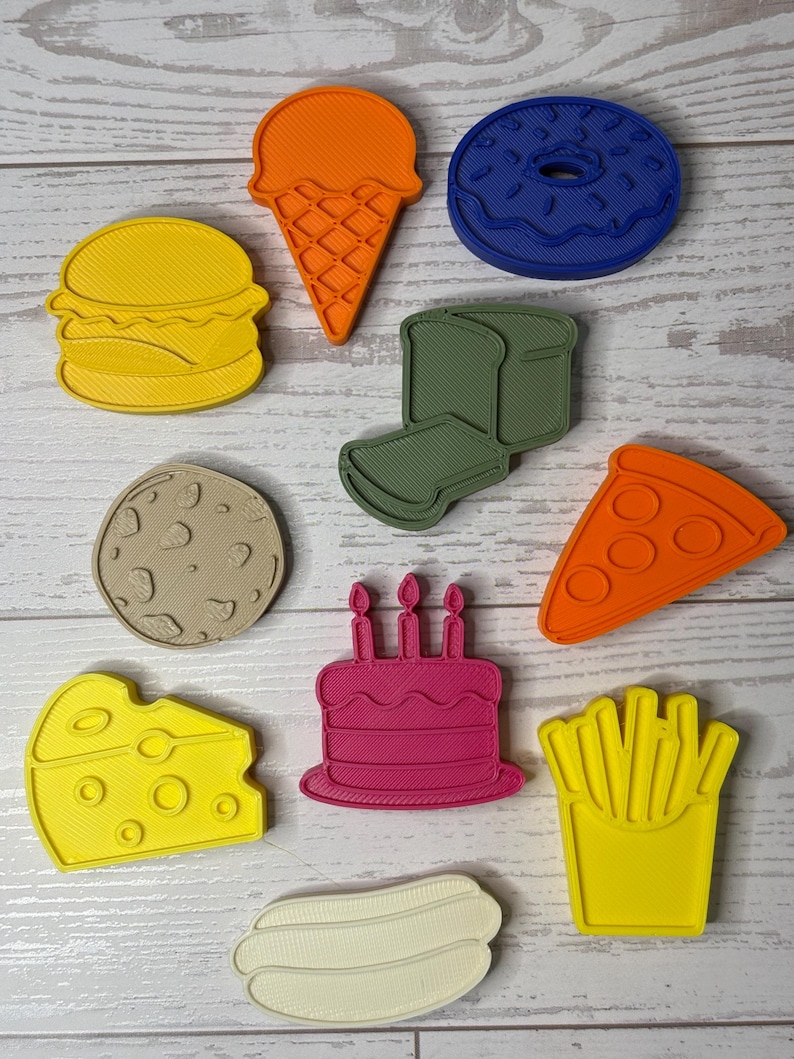 May include: A collection of ten plastic food-themed cookie cutters in various colors, including a yellow burger, orange ice cream cone, blue donut, green sandwich, tan cookie, orange pizza slice, yellow cheese wedge, pink birthday cake, and white hot dog.