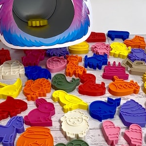 May include: A purple monster-shaped feeding toy with a wide open mouth and a collection of colorful plastic cookie cutters in various shapes including animals, food, and vehicles scattered in front of the toy on a white wooden surface.