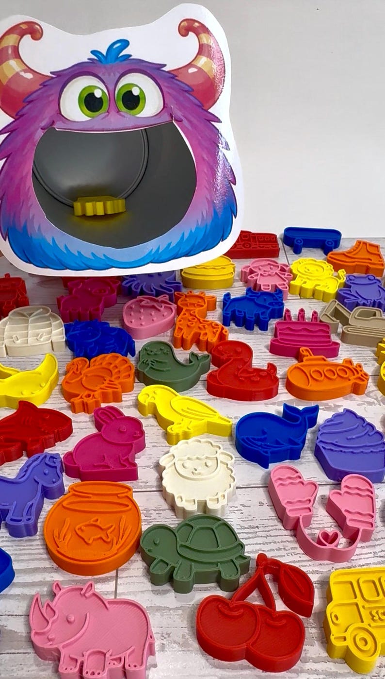 May include: A purple monster-shaped feeding toy with a wide open mouth and a collection of colorful plastic cookie cutters in various shapes including animals, food, and vehicles scattered in front of the toy on a white wooden surface.