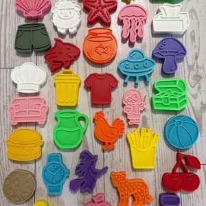 May include: A collection of multi-colored plastic cookie cutters in various shapes including animals, food items, clothing, and household objects. The set features items like a fish, starfish, sheep, jellyfish, shorts, t-shirt, burger, fries, and a cherry.