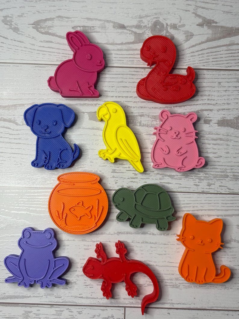May include: A collection of ten colorful plastic animal-shaped cookie cutters including a pink rabbit, red snake, blue dog, yellow parrot, pink hamster, orange fishbowl, green turtle, purple frog, red lizard, and an orange cat.