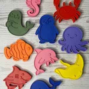May include: A collection of ten plastic cookie cutters in various shapes including a pink seahorse, red crab, green seal, blue penguin, purple octopus, orange fish, pink dolphin, yellow stingray, red shark, and a blue whale, all featuring textured surfaces.