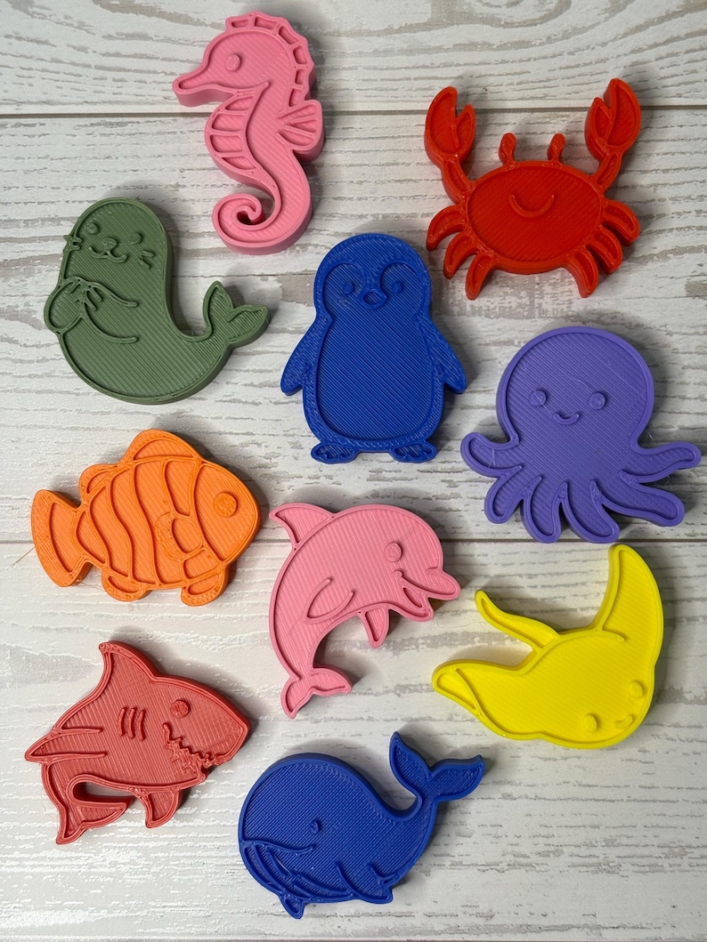 May include: A collection of ten plastic cookie cutters in various shapes including a pink seahorse, red crab, green seal, blue penguin, purple octopus, orange fish, pink dolphin, yellow stingray, red shark, and a blue whale, all featuring textured surfaces.
