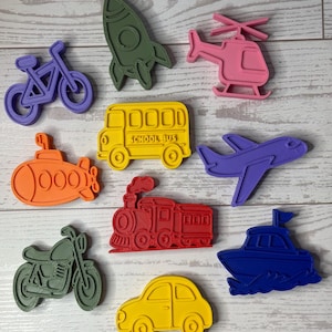 May include: A collection of ten plastic cookie cutters in various shapes including a purple bicycle, olive green rocket, pink helicopter, orange submarine, yellow school bus with text, purple airplane, red steam train, olive green motorcycle, yellow car, and blue boat.