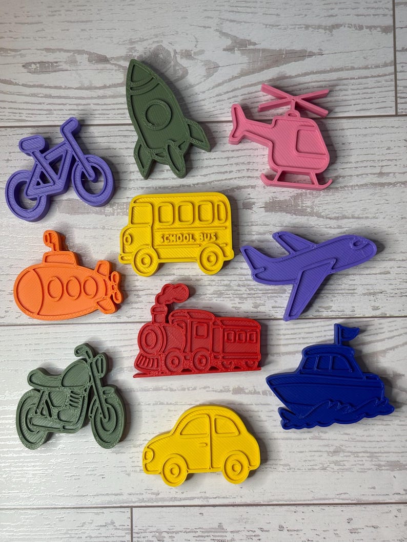 May include: A collection of ten plastic cookie cutters in various shapes including a purple bicycle, olive green rocket, pink helicopter, orange submarine, yellow school bus with text, purple airplane, red steam train, olive green motorcycle, yellow car, and blue boat.
