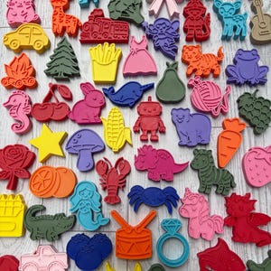 May include: A collection of colorful plastic cookie cutters in various shapes including animals, vehicles, food items, and fantasy characters. The assortment features vibrant red, blue, yellow, pink, green, and orange shapes arranged together.