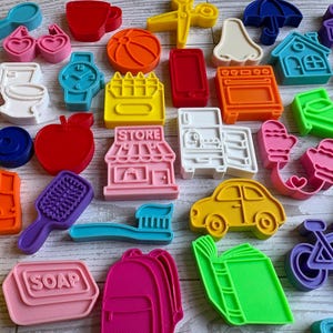 May include: A collection of colorful plastic cookie cutters in various shapes including a store with the word STORE, a soap bar with the word SOAP, a toothbrush, a car, a bicycle, a house, a toilet, a watch, a hairbrush, a boot, and a book.