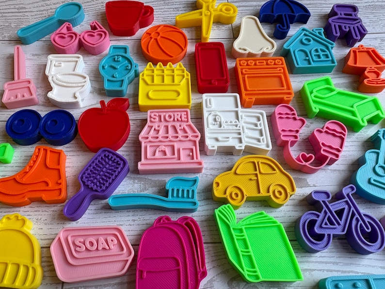 May include: A collection of colorful plastic cookie cutters in various shapes including a store with the word STORE, a soap bar with the word SOAP, a toothbrush, a car, a bicycle, a house, a toilet, a watch, a hairbrush, a boot, and a book.