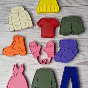 May include: A collection of ten plastic cookie cutters shaped like clothing items, including a yellow beanie, cream puffer jacket, red t-shirt, olive shorts, orange boot, pink mittens, purple socks, pink dress, olive sweater, and blue jeans.