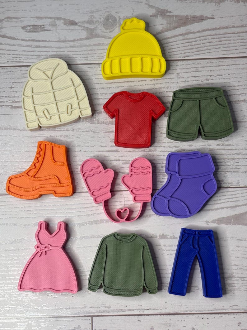 May include: A collection of ten plastic cookie cutters shaped like clothing items, including a yellow beanie, cream puffer jacket, red t-shirt, olive shorts, orange boot, pink mittens, purple socks, pink dress, olive sweater, and blue jeans.
