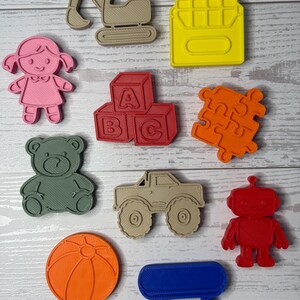 May include: A collection of ten plastic cookie cutters in various shapes including a tan excavator, yellow crayon, pink doll, red alphabet blocks with A, B, and C, orange puzzle piece, green teddy bear, tan monster truck, red robot, orange ball, and blue wagon.