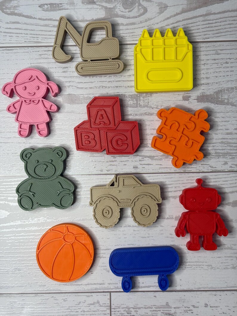May include: A collection of ten plastic cookie cutters in various shapes including a tan excavator, yellow crayon, pink doll, red alphabet blocks with A, B, and C, orange puzzle piece, green teddy bear, tan monster truck, red robot, orange ball, and blue wagon.