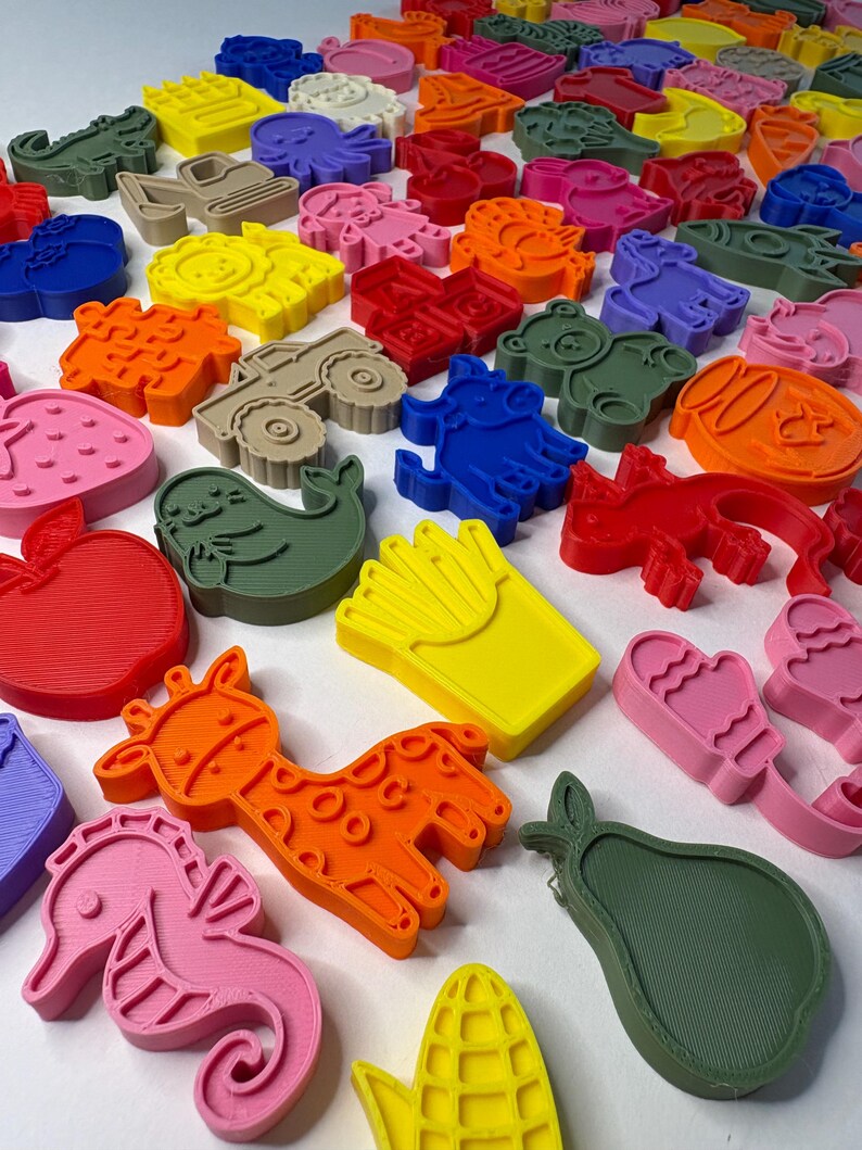 May include: A collection of colorful plastic cookie cutters in various shapes including animals, food items, vehicles, and letters. The assortment features vibrant shades of red, orange, yellow, green, blue, and pink, all arranged together.