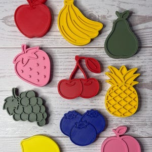 May include: A collection of ten plastic fruit-shaped cookie cutters in various colors, including a red apple, yellow banana, green pear, pink strawberry, red cherries, yellow pineapple, gray grapes, blue blueberries, yellow lemon, and pink peach.