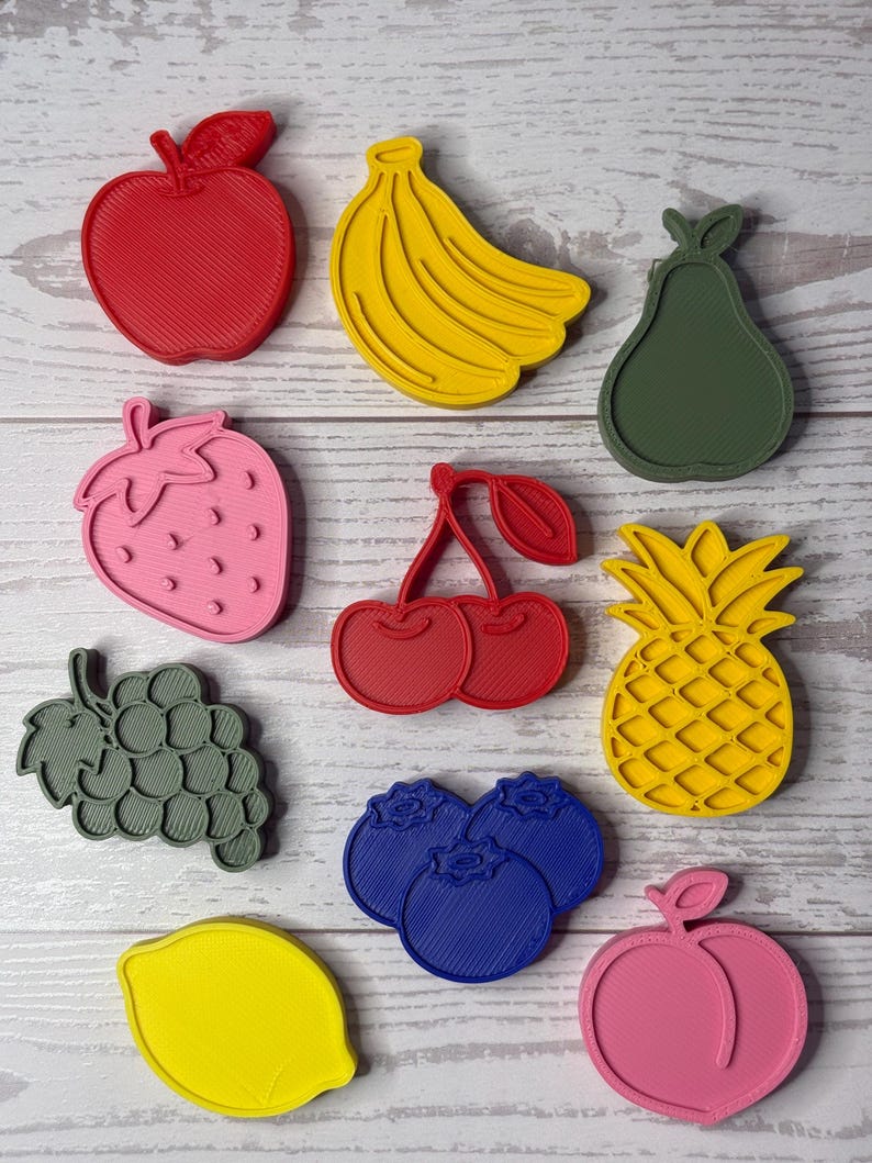 May include: A collection of ten plastic fruit-shaped cookie cutters in various colors, including a red apple, yellow banana, green pear, pink strawberry, red cherries, yellow pineapple, gray grapes, blue blueberries, yellow lemon, and pink peach.