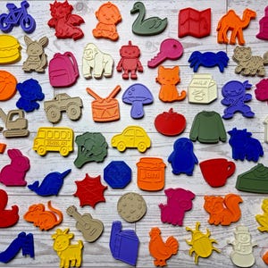May include: A collection of colorful cookie cutters in various shapes, including animals, vehicles, and food items. The cutters are in shades of red, orange, yellow, green, blue, and purple. The shapes include a crab, bicycle, baby, and a school bus.