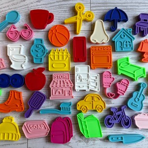 May include: A collection of colorful plastic cookie cutters in various shapes including a spoon, cup, scissors, umbrella, chair, glasses, ball, nose, house, lamp, broom, toilet, watch, crayons, phone, oven, bed, apple, store, fridge, mittens, key, hammer, boot, brush, toothbrush, car, guitar, hat, soap, backpack, book, bicycle, bathtub, and knife.